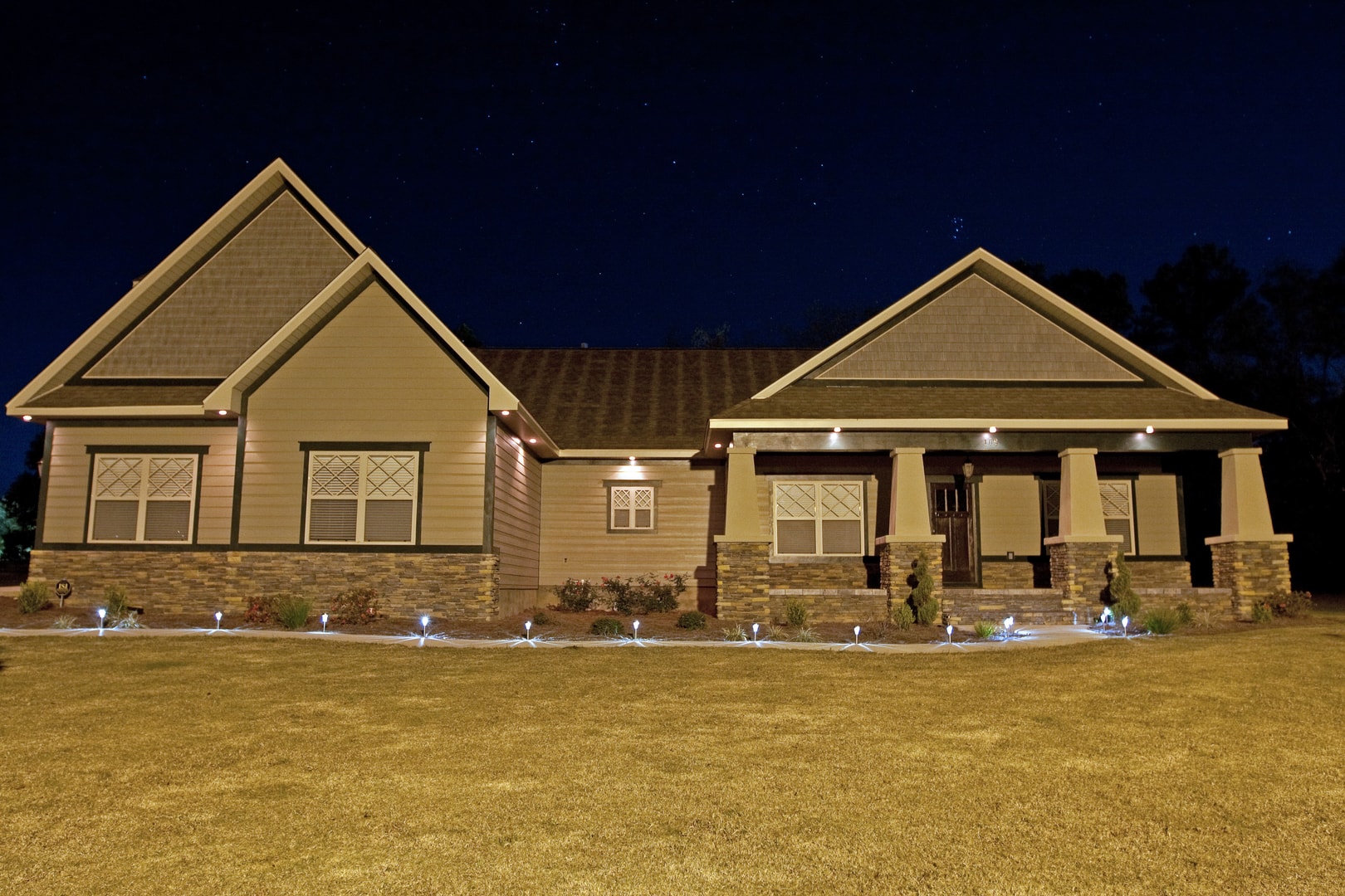Craftsman bungalow at night with landscape lighting, tan siding, stacked stone base and covered porch