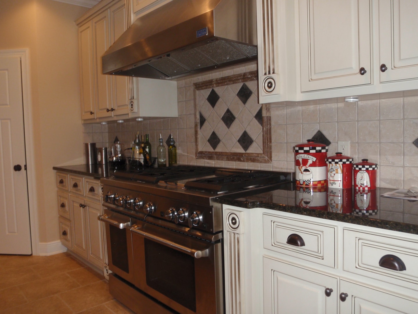 Wide view of kitchen with pro range, diamond tile backsplash, cream glazed cabinets and stainless hood