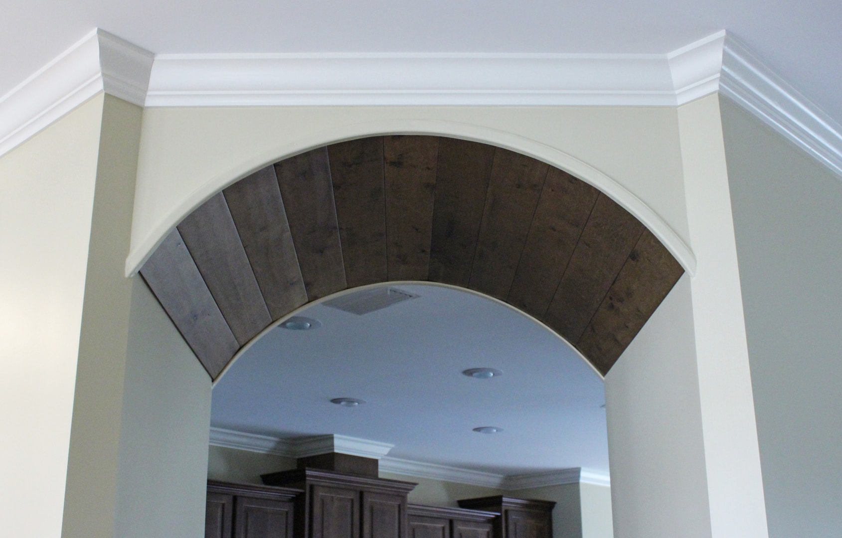 Dramatic arched opening with dark wood plank interior soffit, white crown molding and view into kitchen with dark espresso cabinets