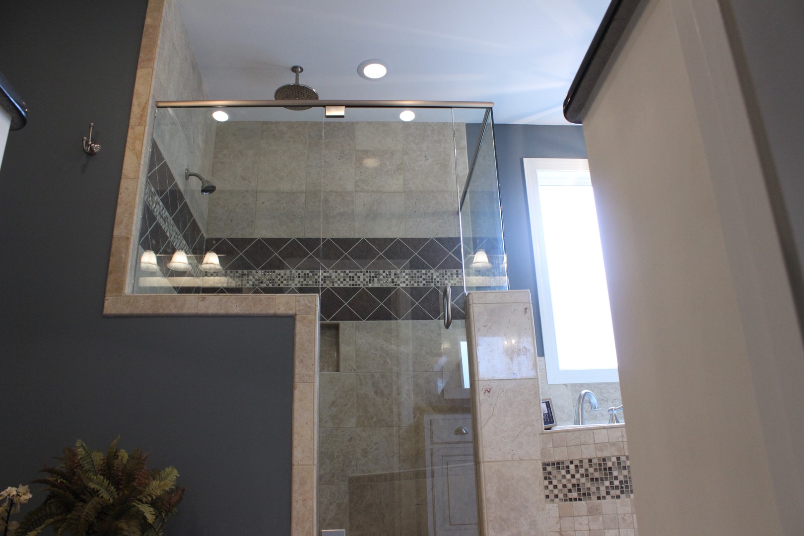 Frameless glass shower enclosure with travertine tile, diamond-set dark tile accent band, mosaic strip, rain head and dark gray walls