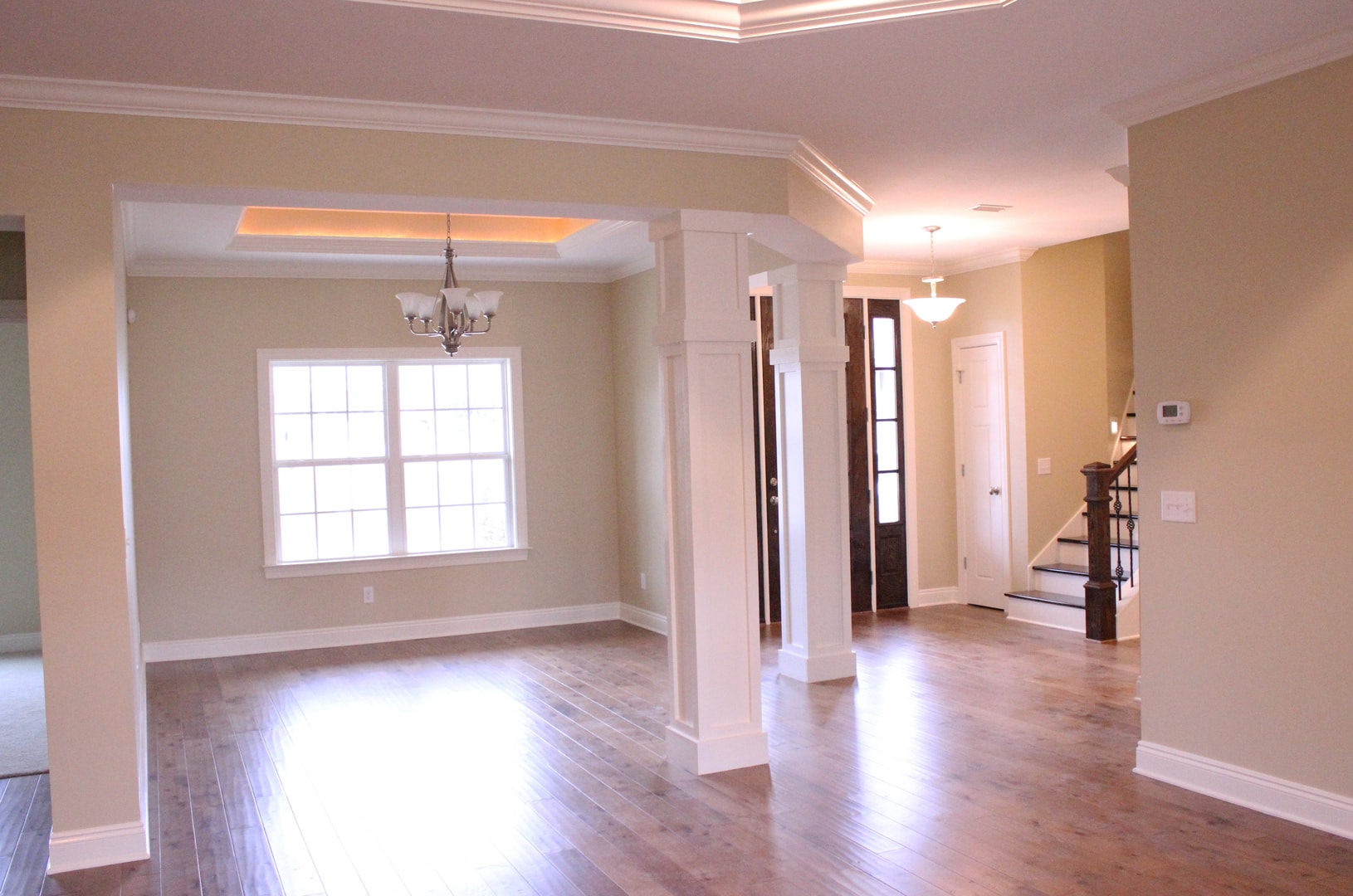 Open plan living and dining with warm hardwood floors, white columns, tray ceiling with cove lighting, chandelier and iron stair railing