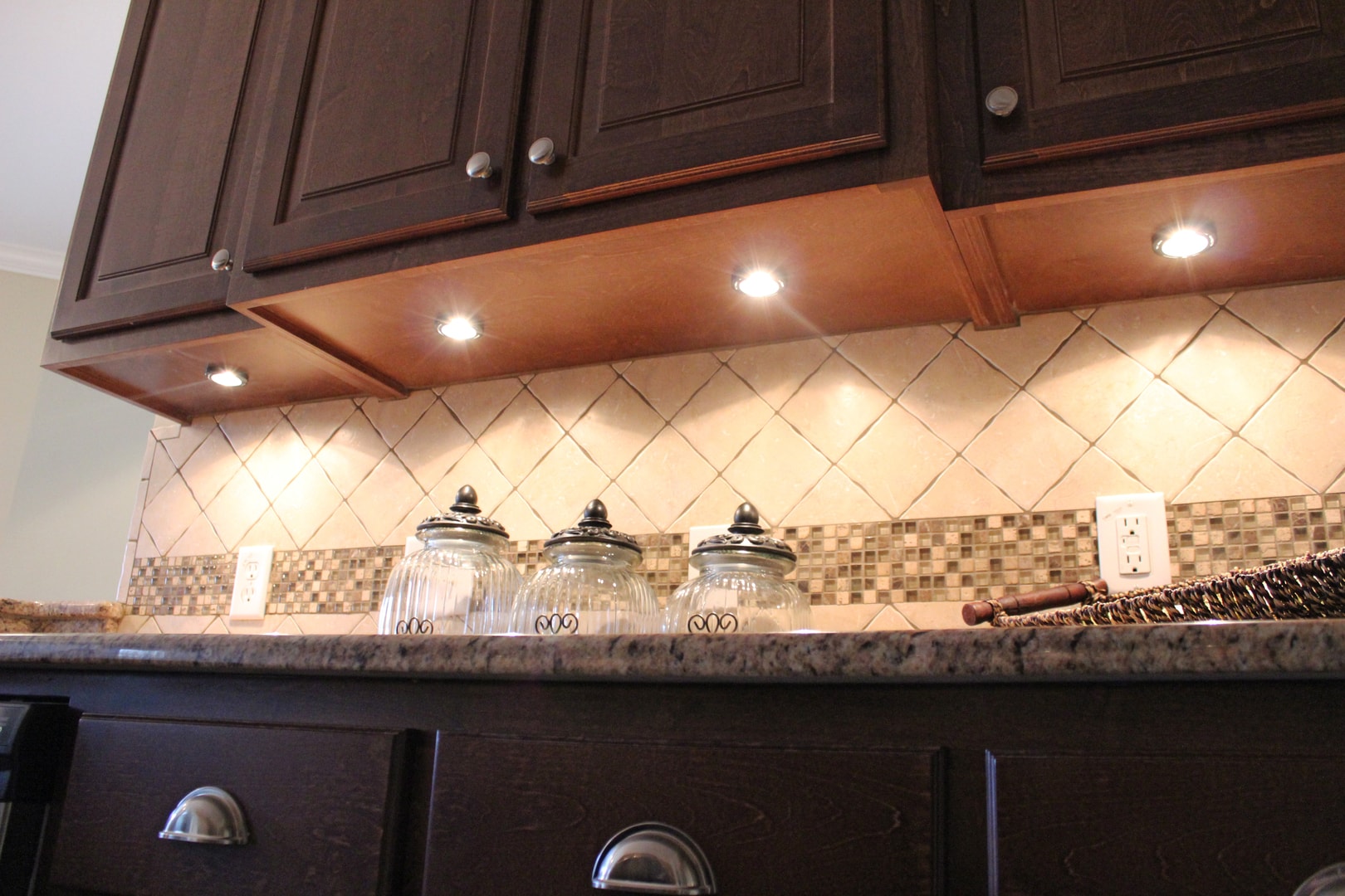 Dark espresso kitchen cabinets with under-cabinet puck lights, diamond tile backsplash with mosaic strip and glass canisters on granite counter