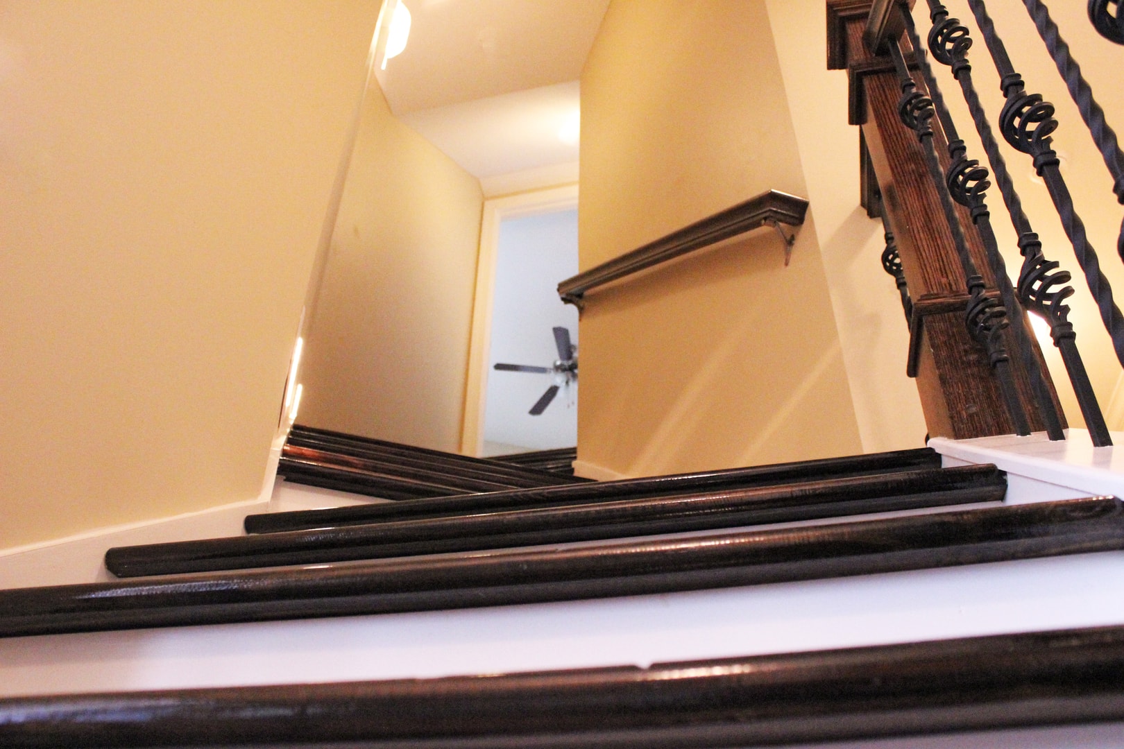 Looking down staircase with dark wood treads, white risers, ornate iron balusters with basket scroll detail and warm yellow walls