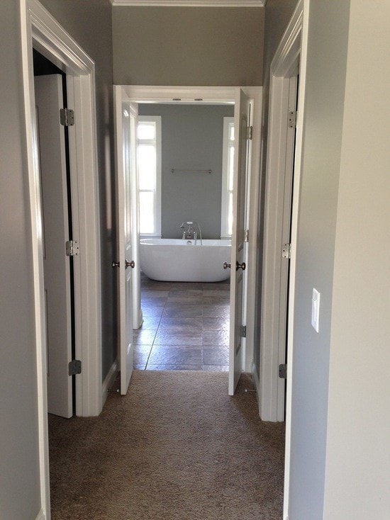 Hallway with gray walls, carpet and white doors with view through to freestanding tub and dark tile master bath