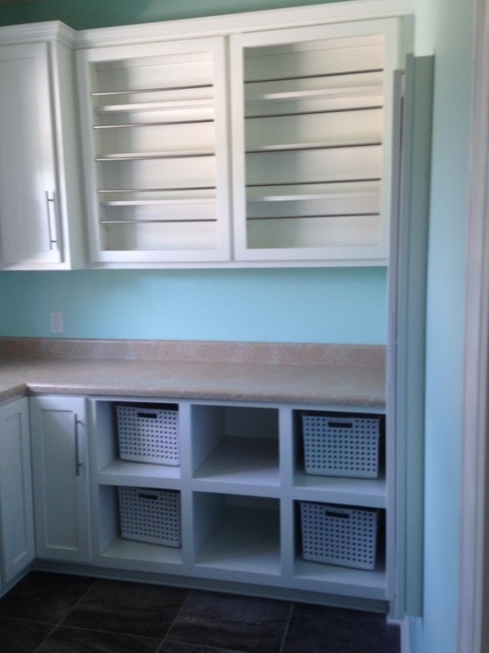 Laundry room with white louvered upper cabinets, teal walls, travertine counter, open cubbies with baskets and dark tile