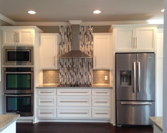 Kitchen appliance wall with white shaker cabinets, double ovens, microwave, stainless fridge and glass tile backsplash