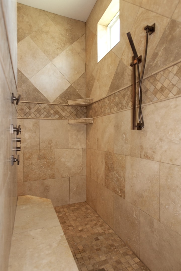 Large-format travertine shower with diamond mosaic border, corner bench and natural light window